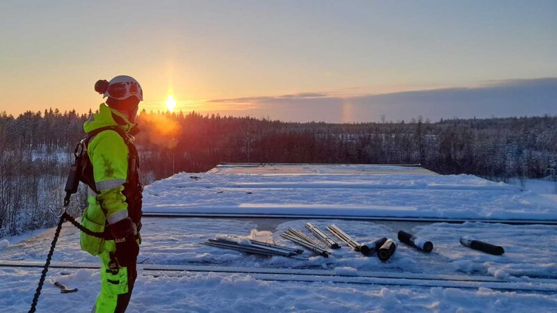 Työvaatteisiin pukeutunut mies seisoo lumisten työmaatilojen katolla ja katsoo horisontissa näkyvää auringonlaskua.