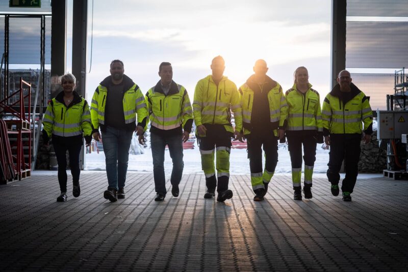 Rivi työvaatteisiin pukeutuneita naisia ja miehiä kävelee kohti kameraa hämärässä hallissa.