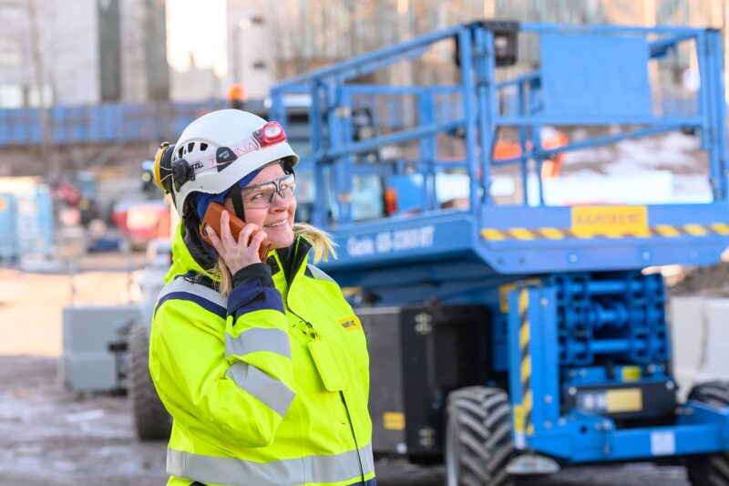 Kypäräpäinen nainen puhuu puhelimeen työmaalla takanaan henkilönostin.