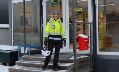 Työvaatepukuinen nainen seisoo vuokraamon ulko-oven edessä ja hymyilee kameralle.