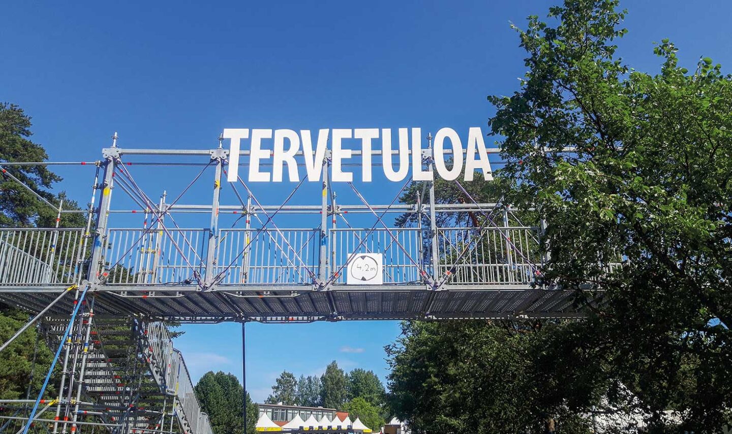 Telineistä koottu tien ylittävä kulkusilta kesäisessä tapahtumassa. Kyltissä lukee Tervetuloa.