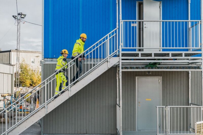 Kaksi henkilöä kipuaa työmaatilojen kulkutietä ylöspäin kohti raollaan olevaa ovea.