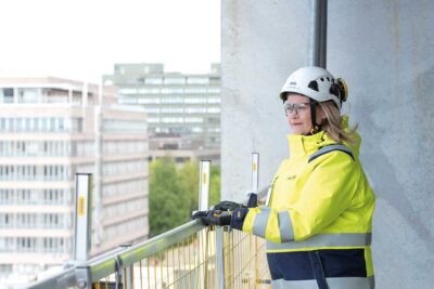 Kypäräpäinen huomiotakkiin pukeutunut nainen seisoo keskeneräisellä parvekkeella kaiteen takana ja katsoo yli kaupungin.