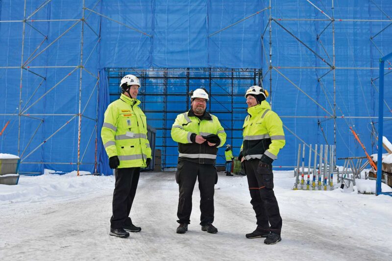 Kolme miestä talvisissa työmaavaatteissa työmaan sääsuojan edessä