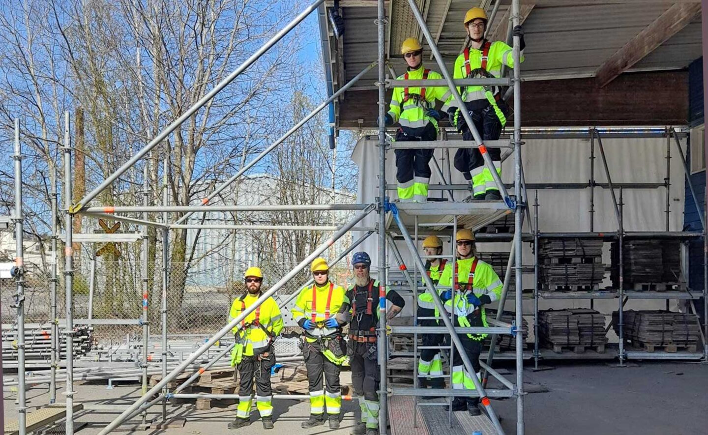 Neljä työvaatepukuista miestä seisoo rakennustelineen tasoilla ja kolme seisoo maassa telineen vierellä.